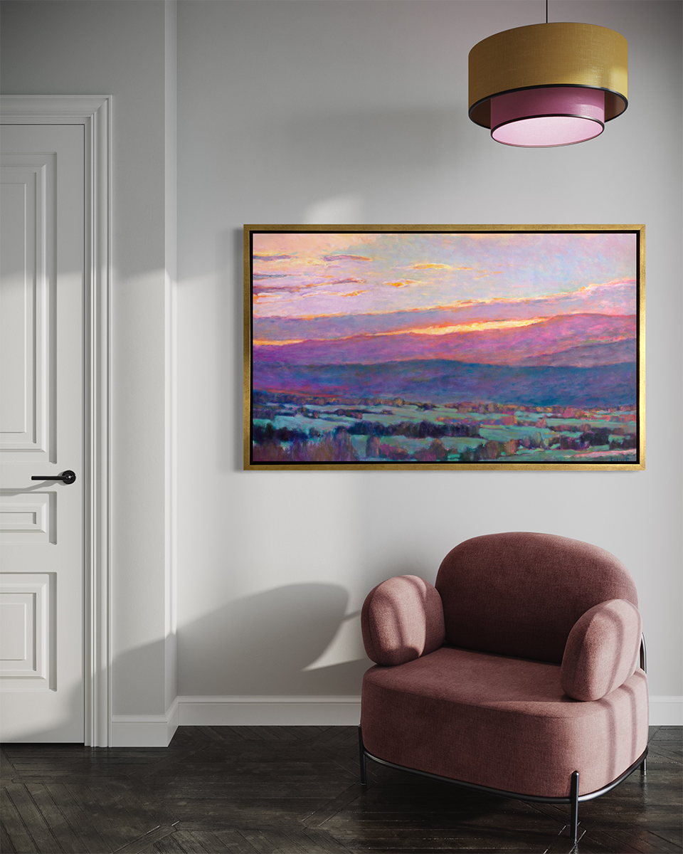 A pink, blue and green abstract landscape print in a gold floater frame hangs on a white wall above a soft mauve armchair with a yellow and pink pendant light hanging from above.