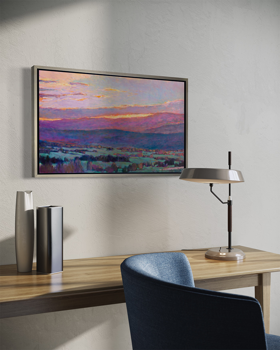 A side view of a pink, blue and green abstract landscape print in a silver floater frame hangs on a white wall above a natural wood desk with a small desk lamp, two vases and a blue desk chair sits in the foreground.