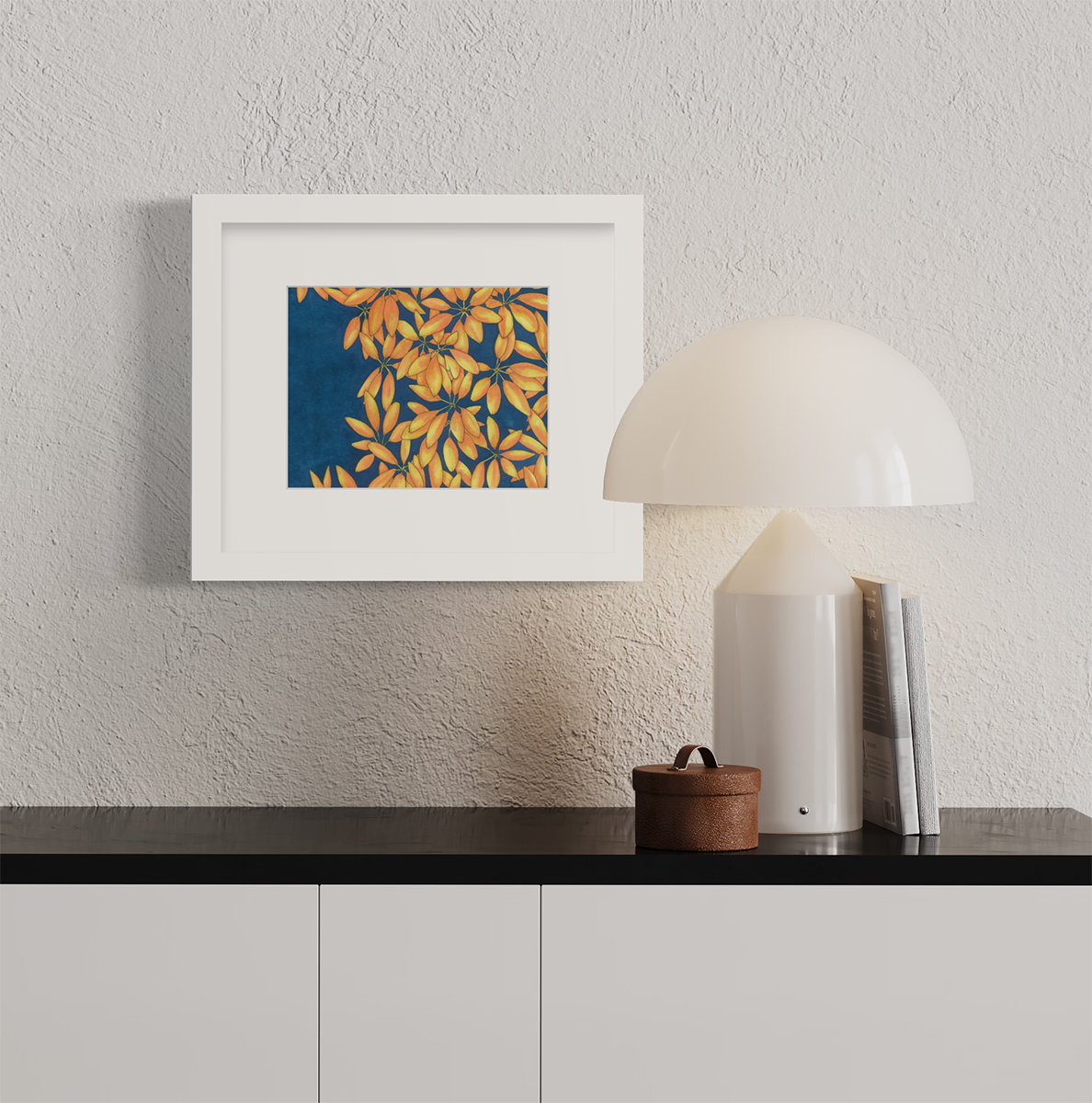 A blue, yellow and orange plant print in a white frame with a mat hangs on a white textured wall above a black surface with a white lamp, two books and a small brown container.