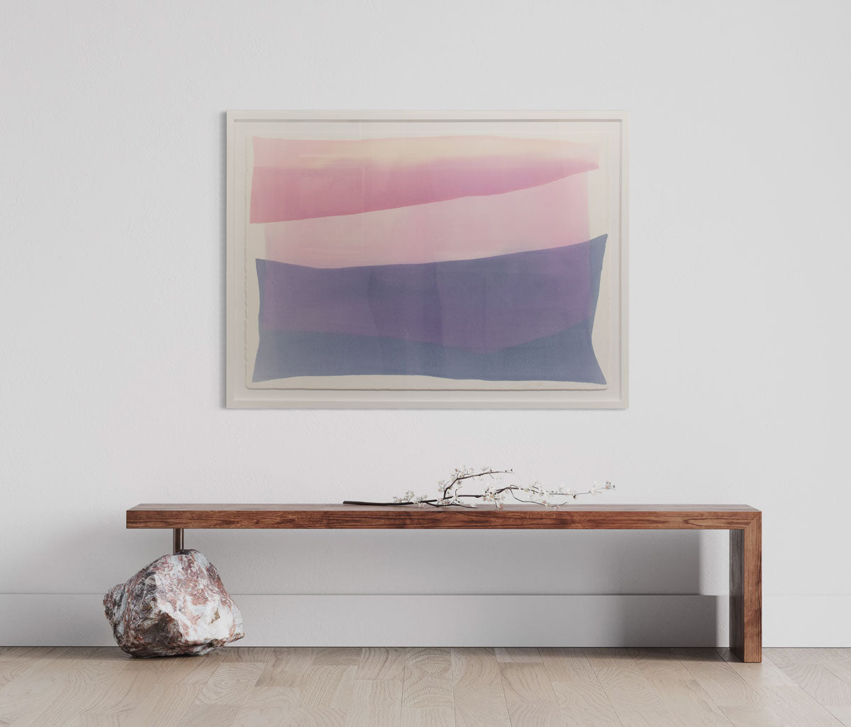A framed pink and violet abstract watercolor painting hangs on a white wall above a modern wooden bench with a natural rock as one foot of the bench. 
