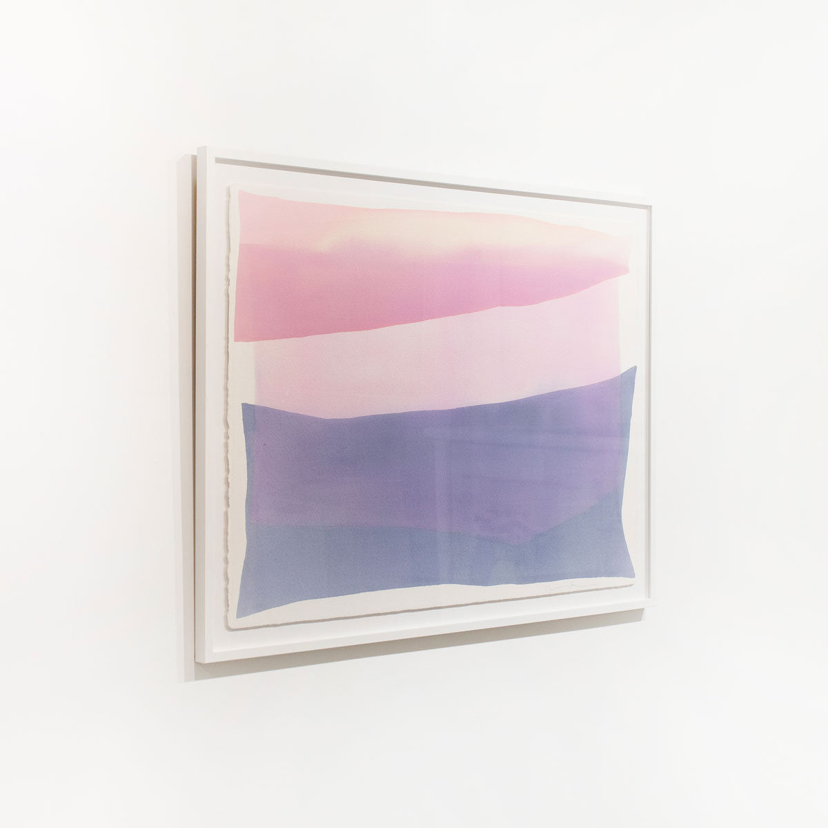 A pink and violet abstract watercolor painting framed in a white frame hangs on a white wall.