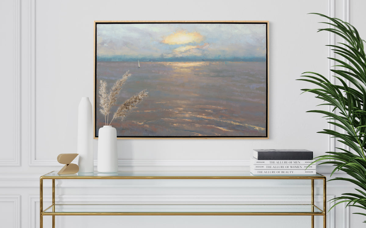 A lavender, orange, yellow and light blue coastal seascape painting by Molly Doe Wensberg hangs in a light wood floater frame on a white wall above a gold trimmed console table with a stack of books, two white vases and a small decorative accent.