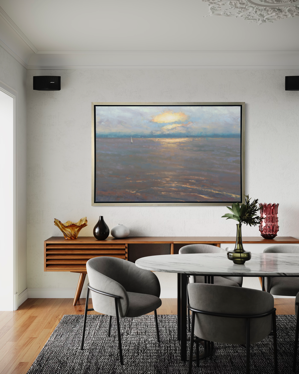 A coastal seascape print in a silver floater frame hangs on a white wall above a stained wood credenza with various colored vases and a white marble dining table with five grey chairs.
