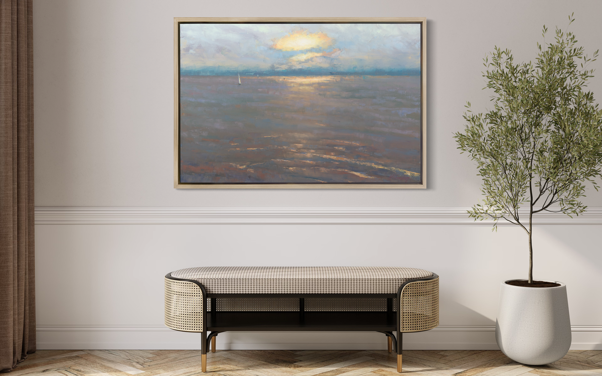 A coastal seascape print in a white oak wood floater frame hangs on a white wall above a rattan bench with a cushion and a tall potted floor plant.
