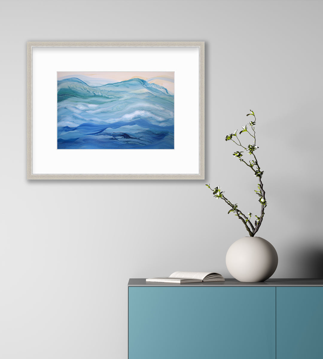 A blue, teal and light orange abstract print in a silver frame with a mat hangs on a white wall above a teal cabinet with an open book and a white rounded vase with greenery stems.