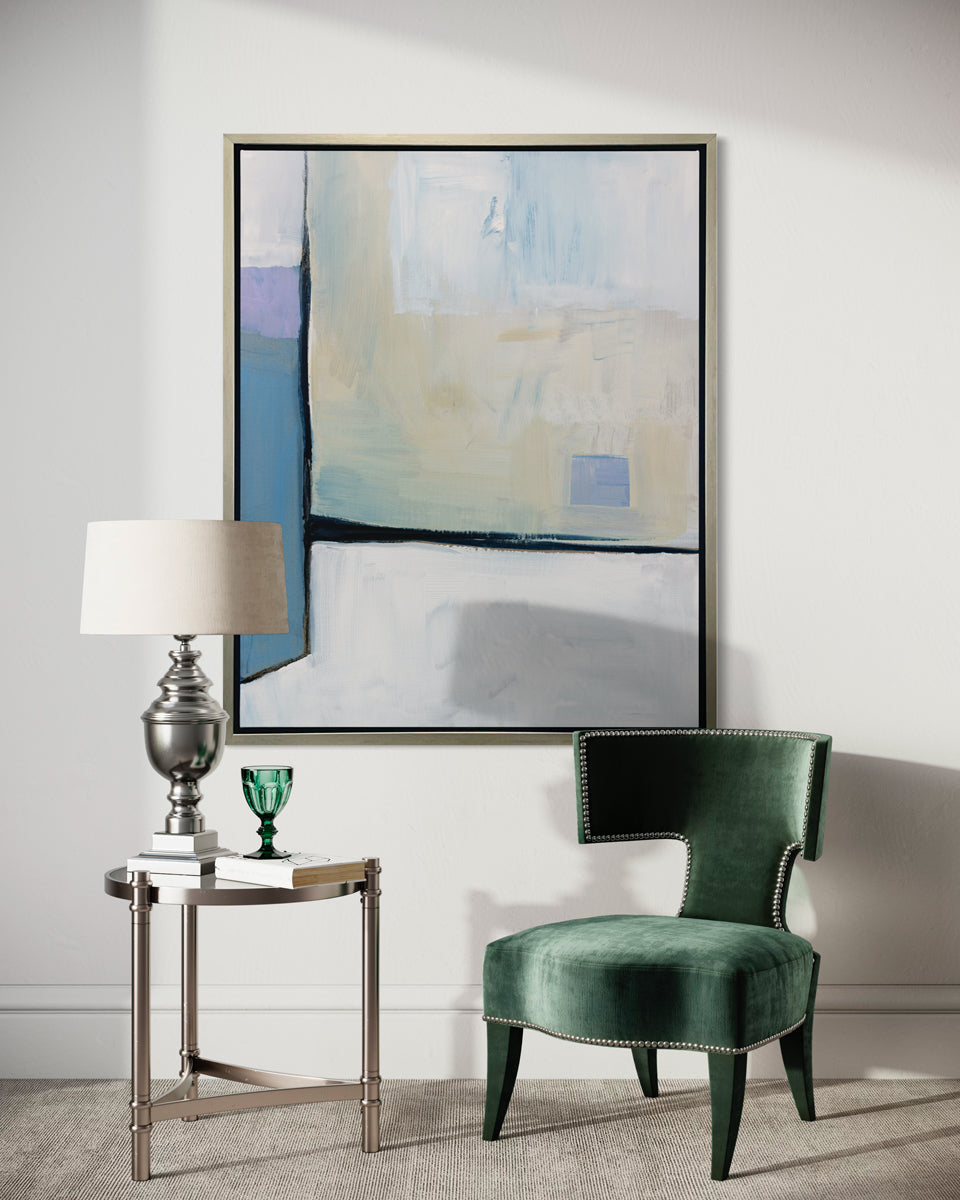 A white, black, blue and chartreuse abstract print in a silver floater frame hangs on a white wall above a nail studded green chair and a round chrome side table with a table lamp and a green glass.