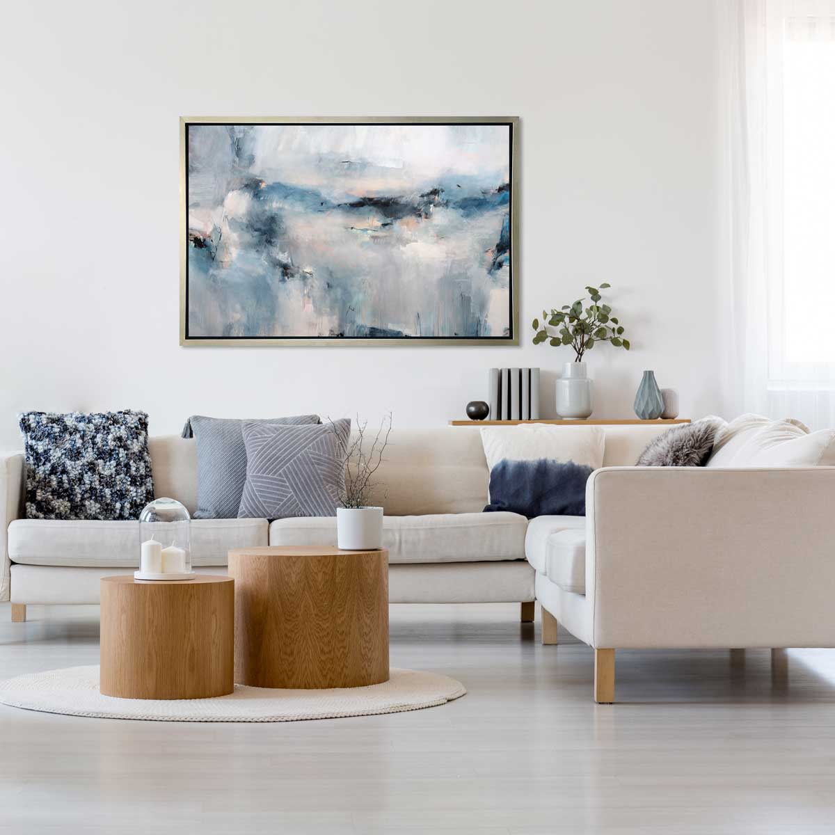 A blue and white abstract print in a silver floater frame hangs on a white wall above a white sectional sofa with blue pillows and two round natural wood coffee tables.