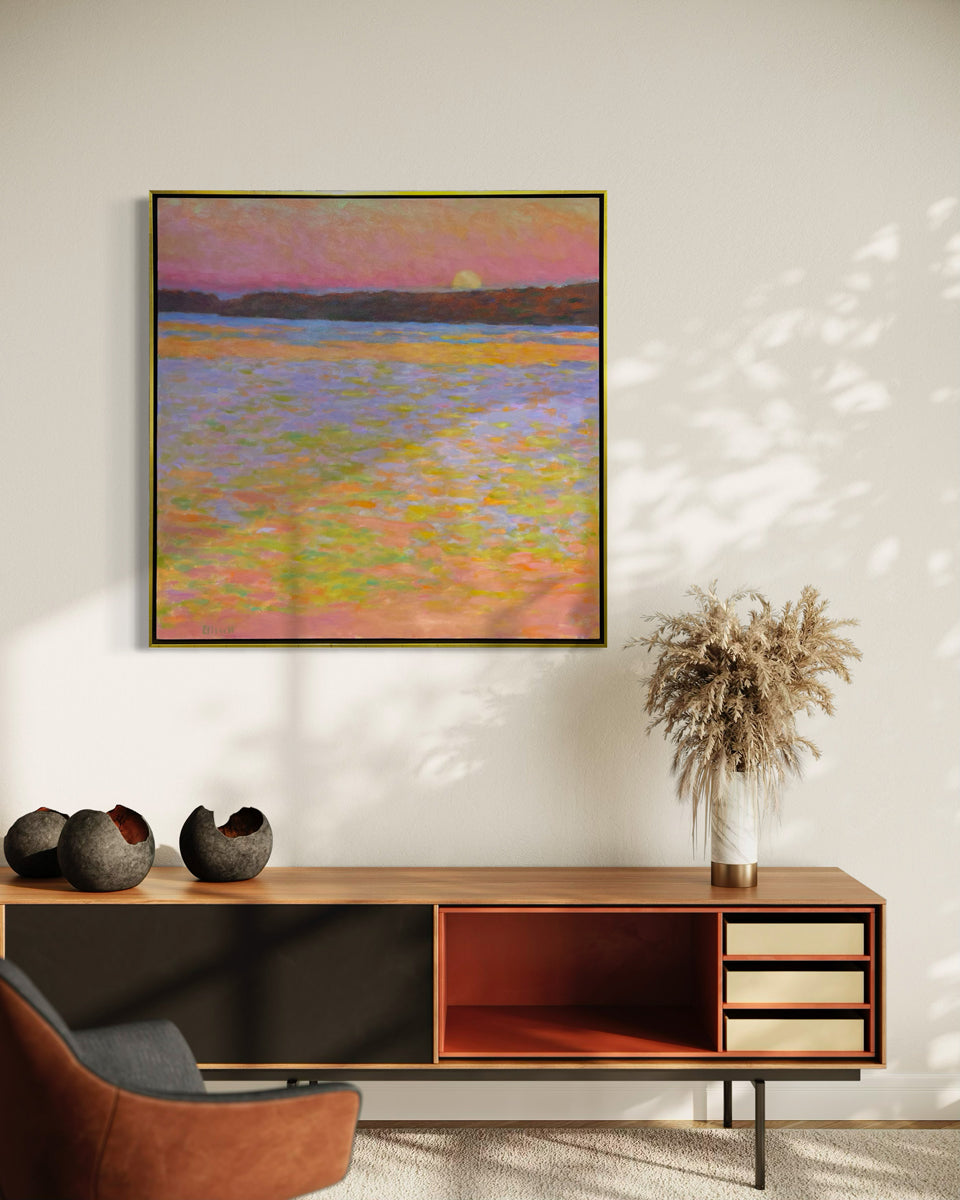 Framed painting of a sunset over water with a colorful sky hanging over a cherry credenza with decorative items