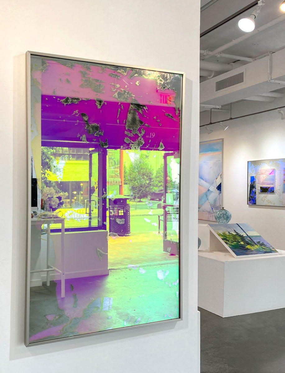A pink fine art mirror hangs on a white column in an art gallery with paintings behind it. 