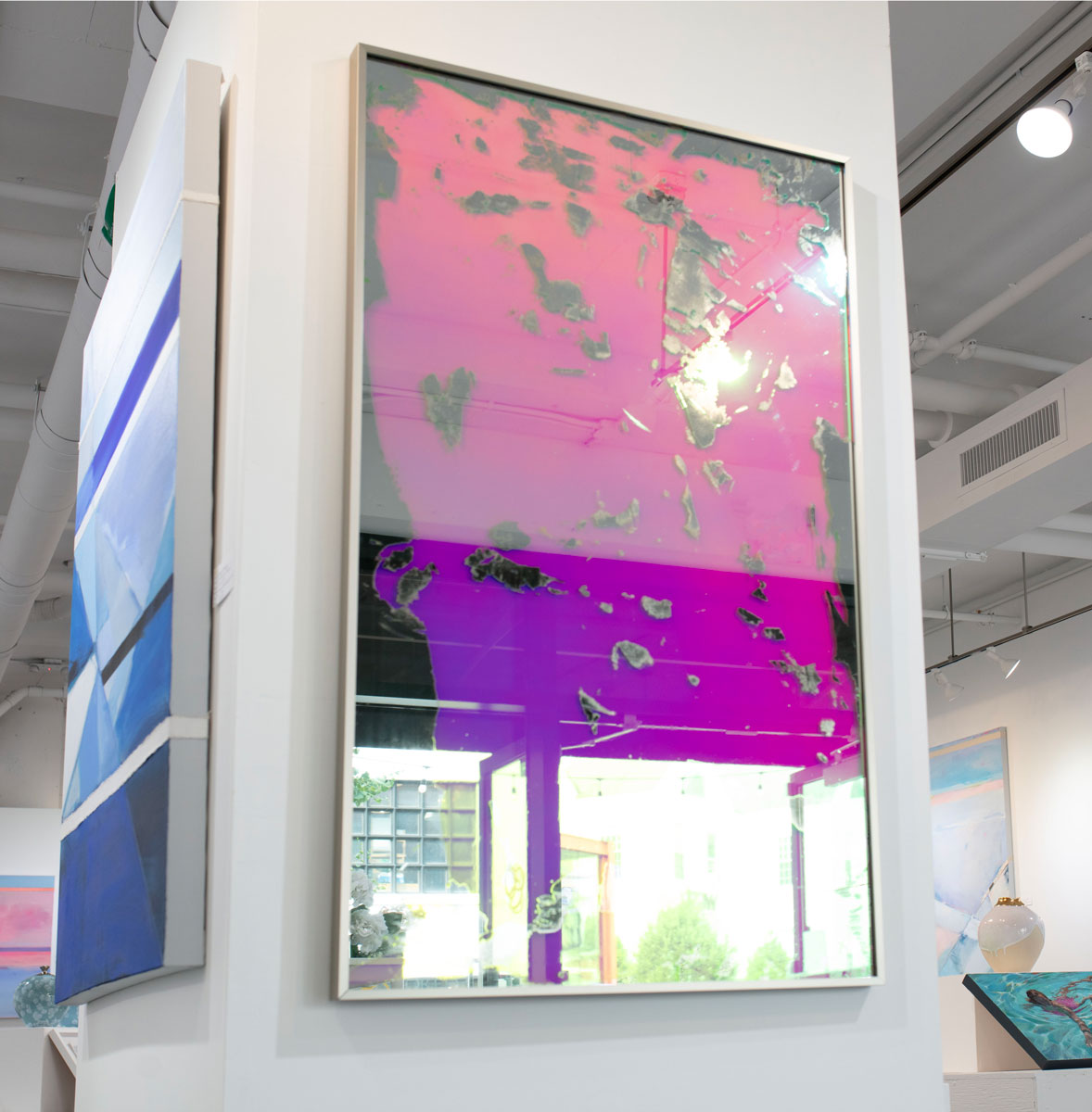 A pink fine art mirror hangs on a column in an art gallery next to a blue abstract painting. 