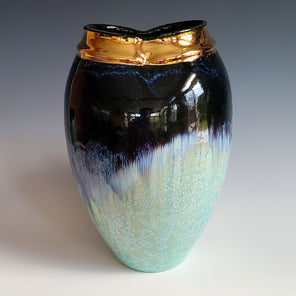 A deep blue, mint green, and gold ceramic vessel rests in front of a dark blue-grey backdrop. 
