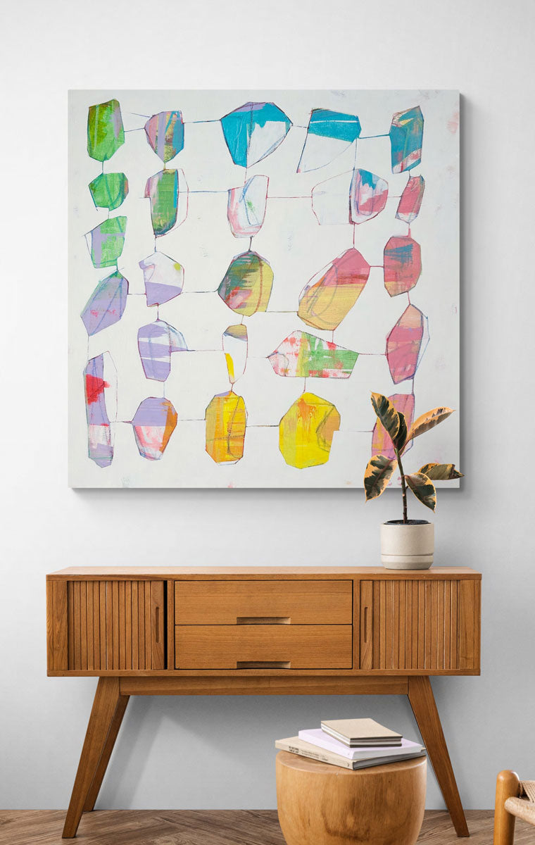 An abstract painting hangs on a white wall above a wooden console table with a potted plant and a wooden table with books. 