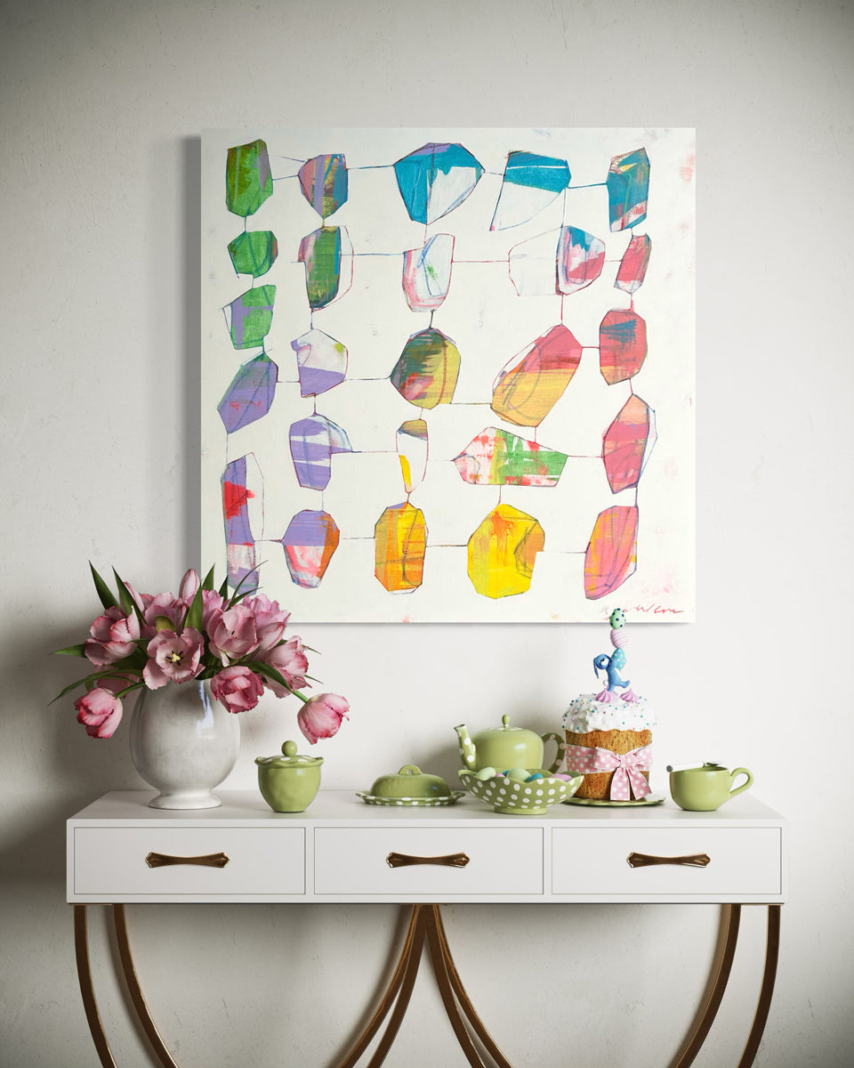 An abstract painting hangs on a white wall above a white modern console table with green and white party decor and a small cake. 