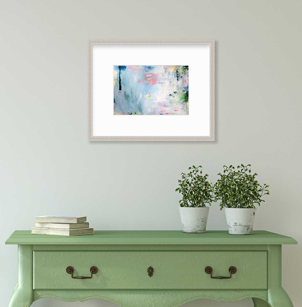 A blue abstract print by Kelly Rossetti in a silver frame with a mat hangs on a white wall above a green console table with a stack of books and two potted plants.