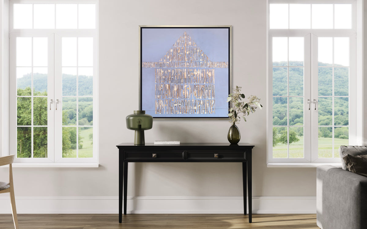 A lavender and beige abstract house print in a silver floater frame hangs on a white wall in between two windows and above a black console table with a green table lamp and a green glass vase with floral stems.