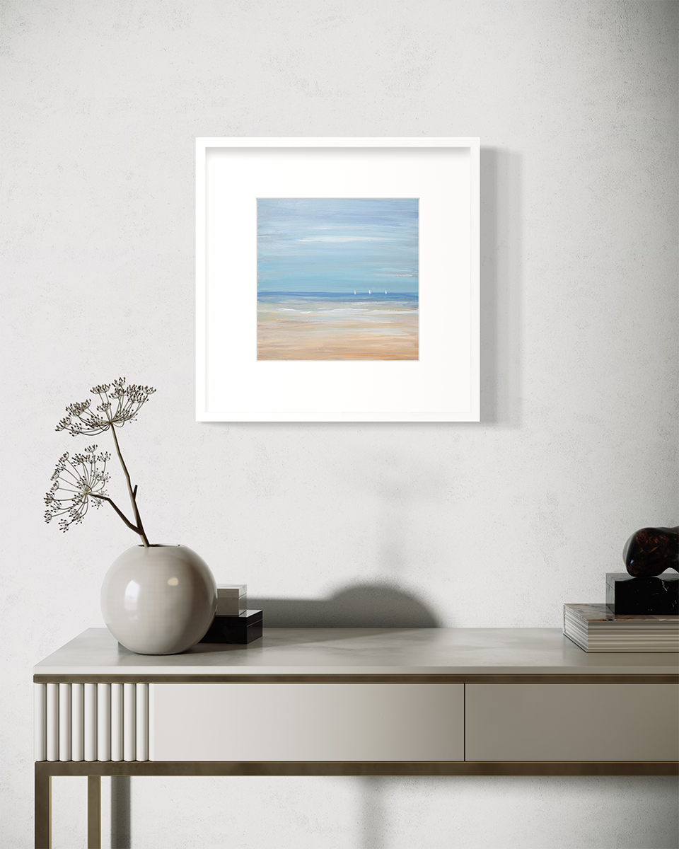 A blue, beige and white abstract seascape print with three small sailboats in a white frame with a mat hangs on a white wall above a white table with a muted grey round vase with decorative stems.