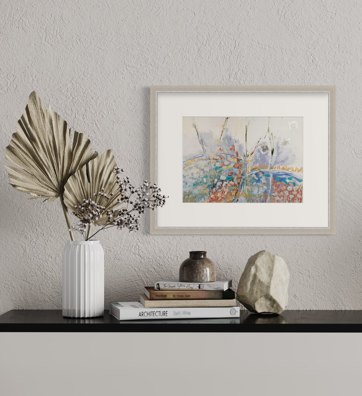 A multicolored abstract floral print in a silver frame with a mat hangs on a white wall above a black surface with a stack of books, a rock and a white vase with decorative stems.