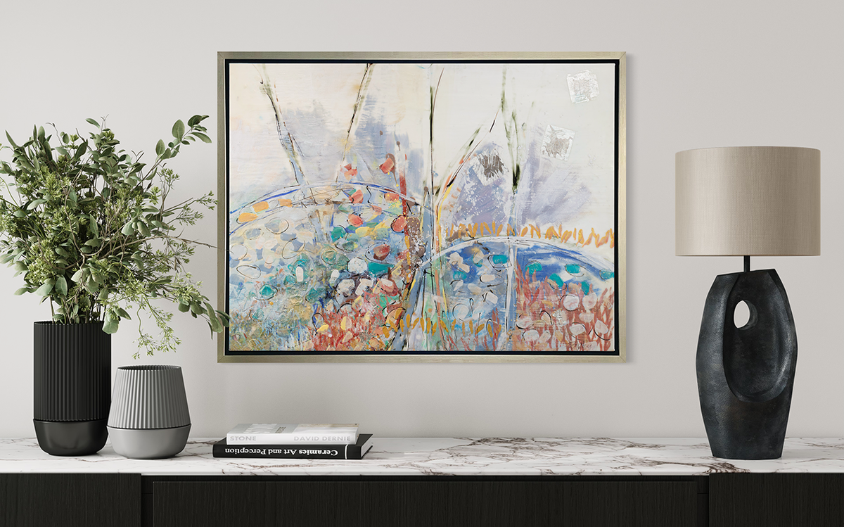 A multicolored abstract floral print in a silver floater frame hangs on a white wall above a white marble surface with a black lamp, a stack of books and two vases with greenery stems.