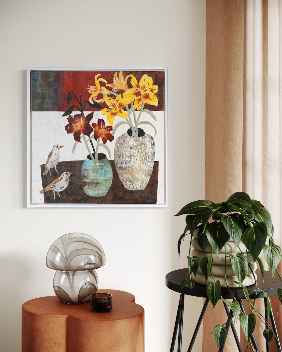 A framed mixed-media still life painting featuring yellow and orange lilies in textured vases, hung on a beige wall in a styled room with a plant and decorative objects.