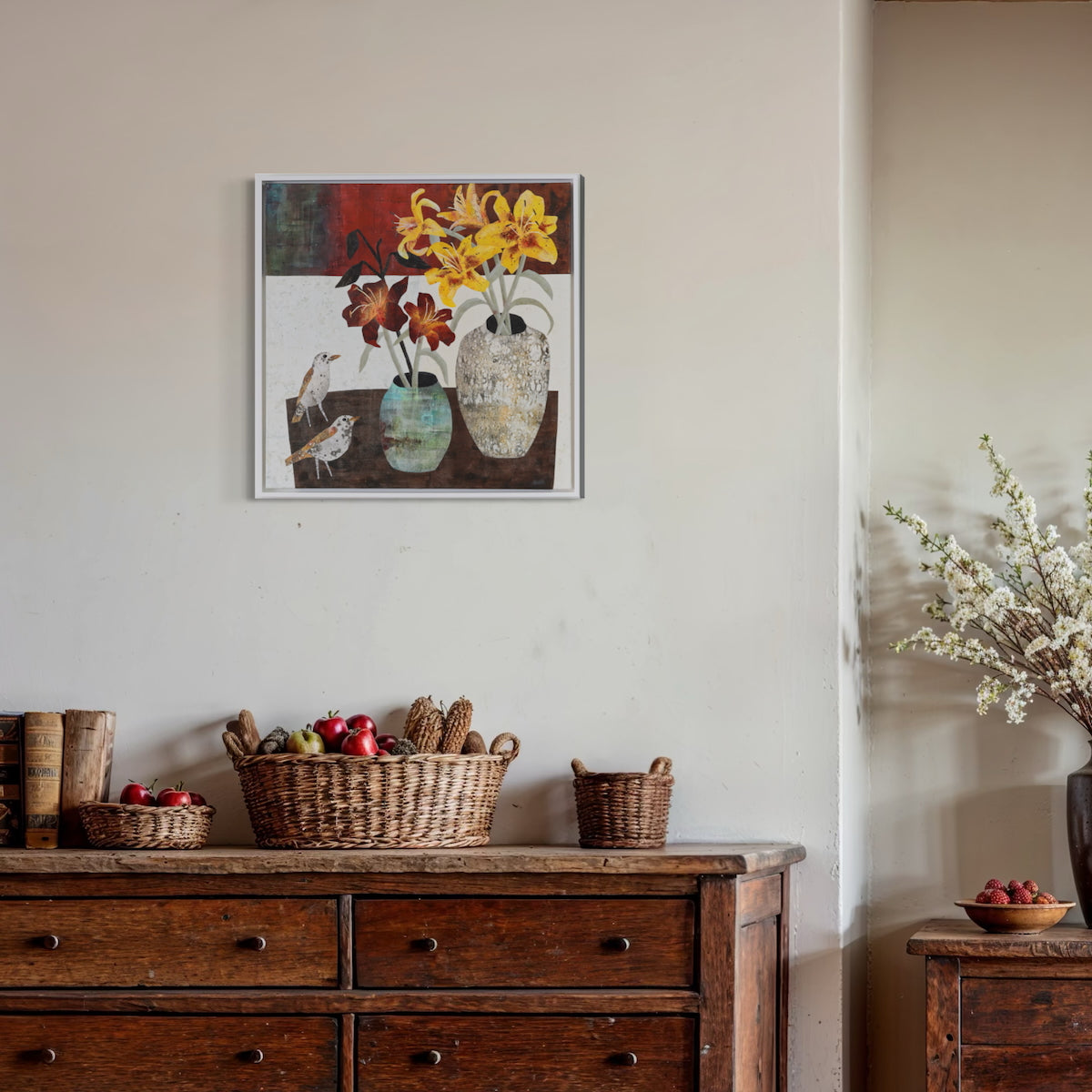 A framed still life painting of yellow flowers and birds in vases, displayed on a beige wall above a rustic wooden dresser with woven baskets and fruit.
