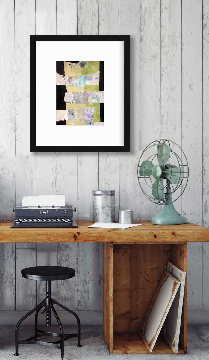 A green, black and beige abstract print by Christine Averill-Green in a black frame with a mat hangs on a grey planked wall above a wood desk with a typewriter, a teal tabletop fan and two silver containers.