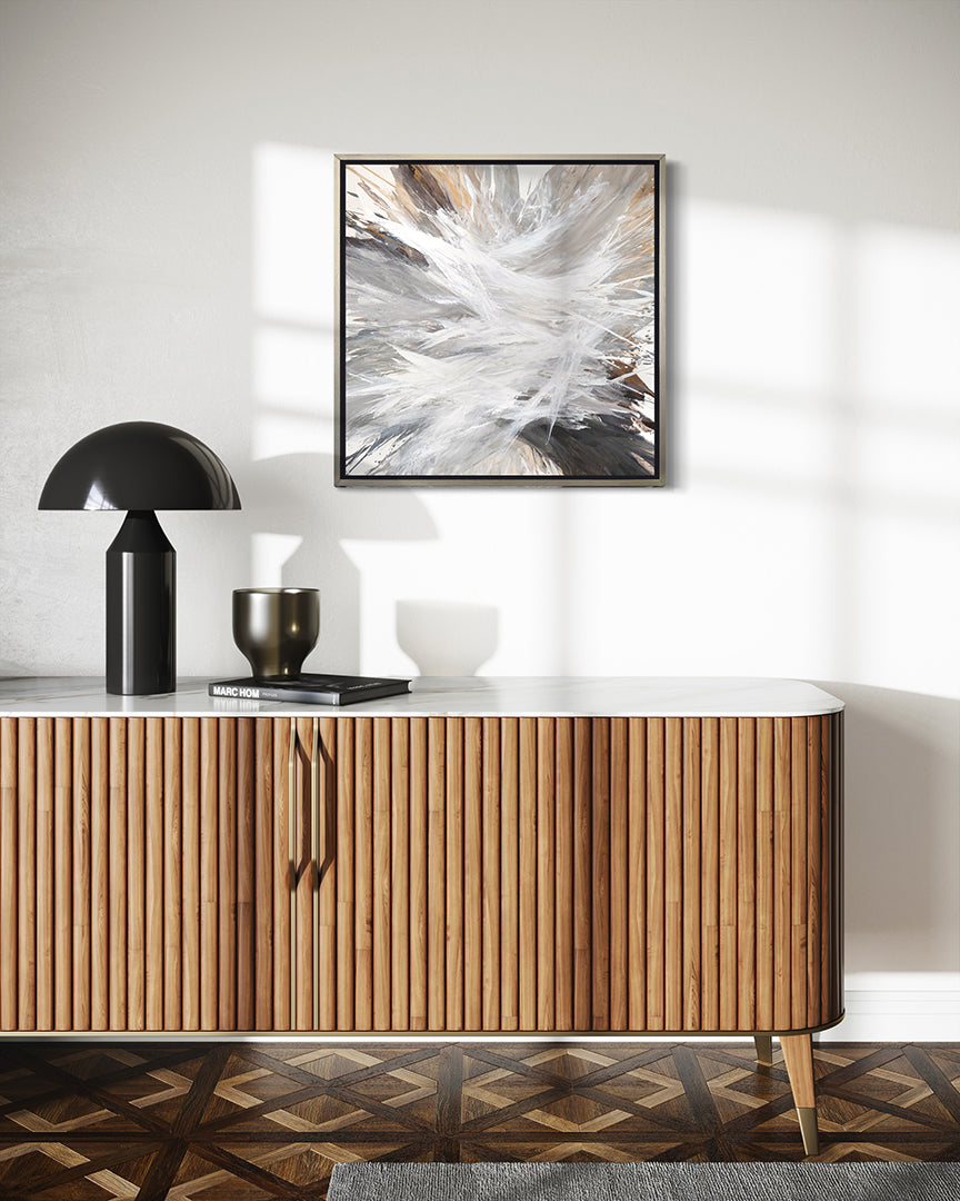 A white, umber, black, grey, yellow ocher and gold abstract print in a silver floater frame hanging on a wall  over a midcentury TV cabinet with a black lamp, vase and book.