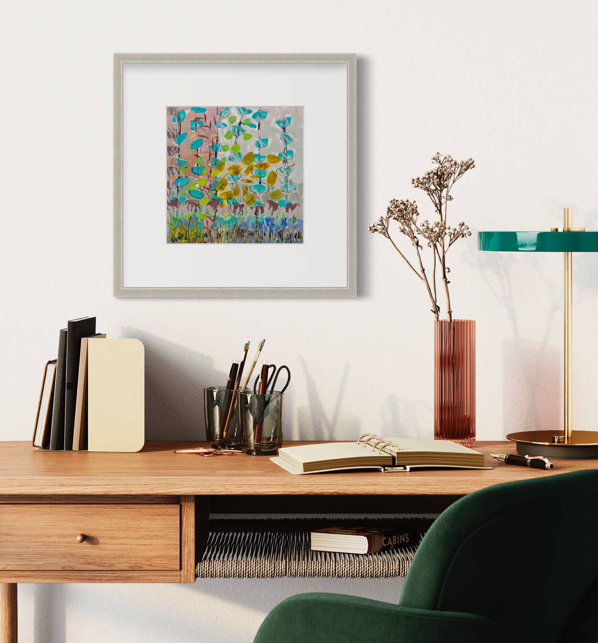 An abstract floral print in a silver frame with a mat hangs on a white wall above a natural wood desk with a small lamp, an open book, a stack of books and a vase with floral stems.