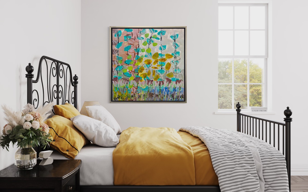An abstract floral print in a silver floater frame hangs on a white wall above a bed with wrought iron headboard and footboard, yellow-orange bedding and a side table with a floral arrangement.