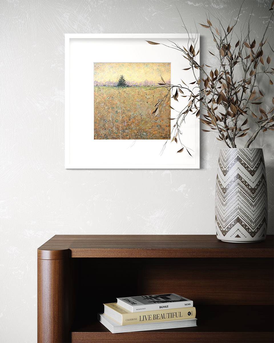 A green, orange and yellow abstract landscape print in a white frame with a mat hangs on a white wall above a dark wood bookshelf with a stack of books and a white and black vase with leaf stems.