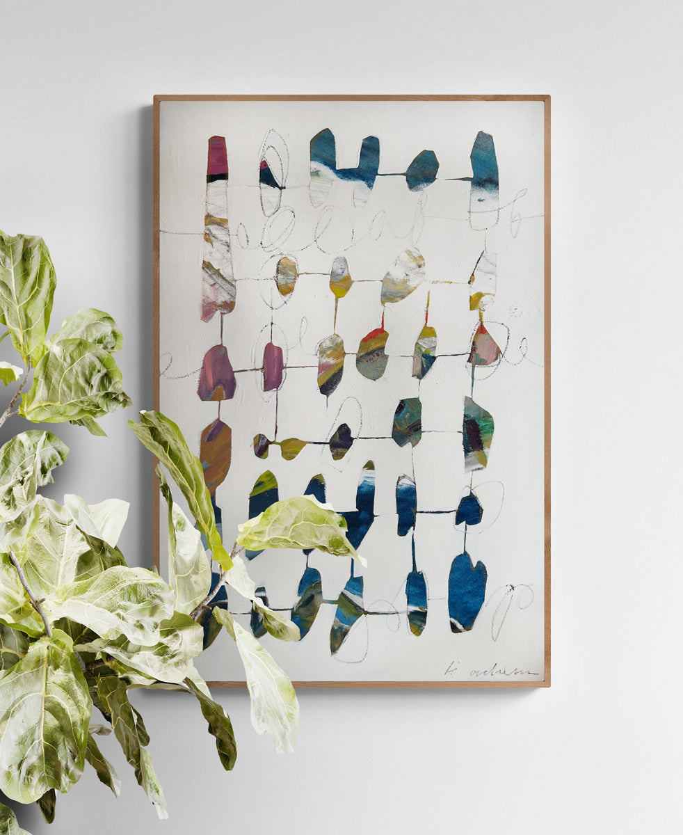 Abstract art piece with colorful geometric shapes in a wooden frame, placed on a light gray wall with a plant to the left.