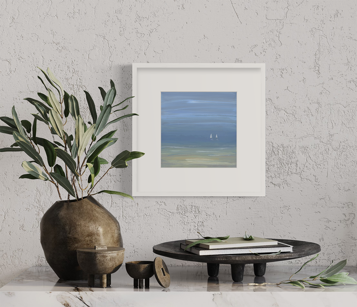 A blue, beige and white abstract seascape print with two small sailboats in a white frame with a mat hangs on an off-white wall above a white marble surface with a wood vase with greenery stems and a tray with books.