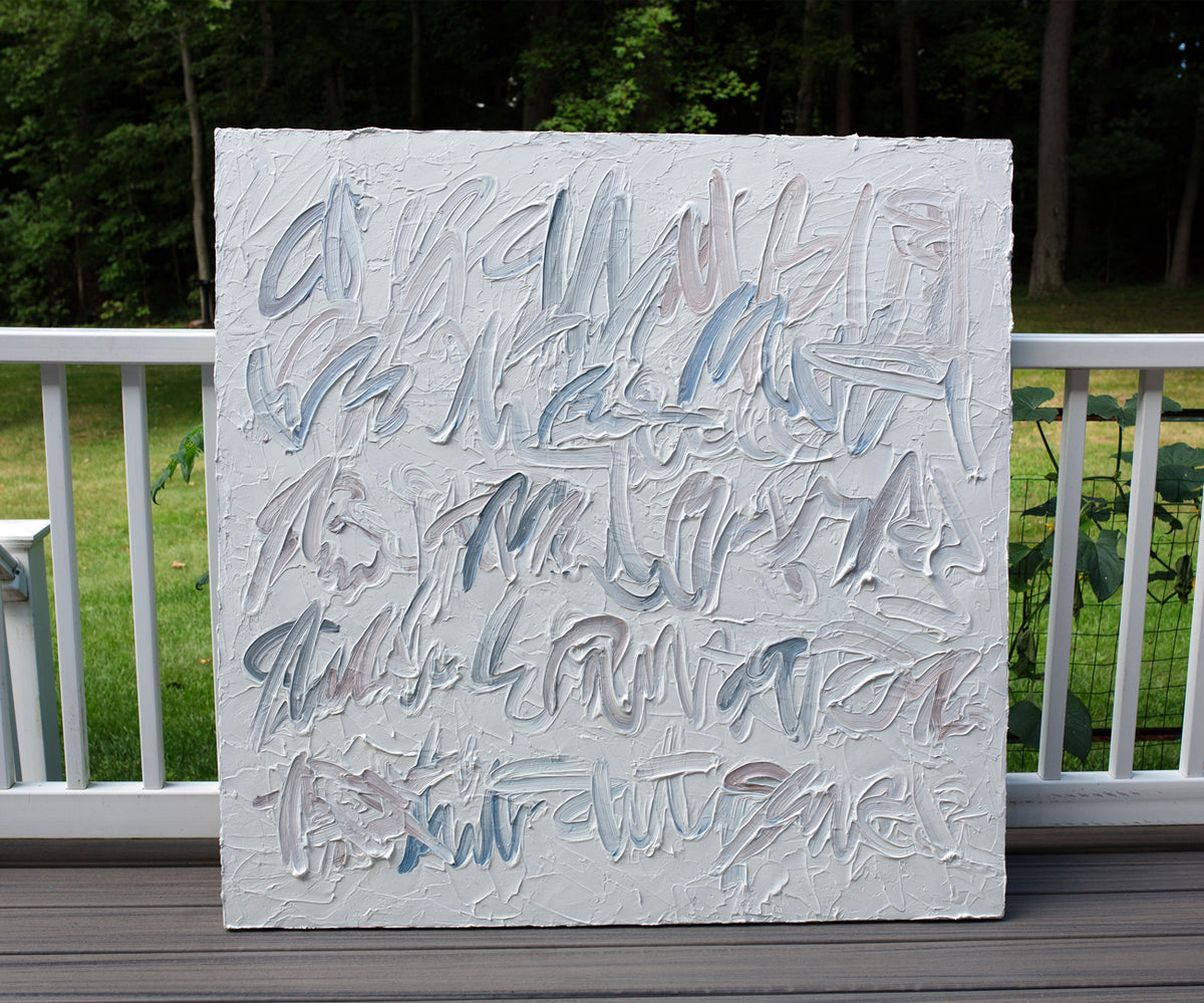 A square canvas painting with thick, textured impasto brushstrokes in shades of gray, blue, and beige, leaning against a white wooden railing on a deck with a green forest background.
