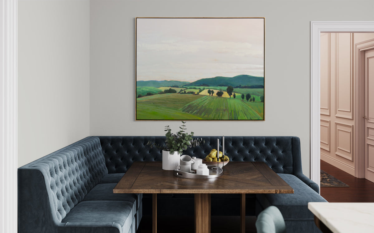 A taupe, yellow and green landscape painting by Molly Doe Wensberg hangs in a light wood floater frame on a white wall above a slate blue tufted corner booth, a dark wood square table and place settings.