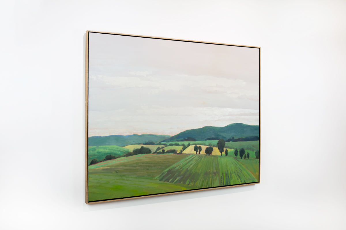 A side view of a taupe, yellow and green landscape painting by Molly Doe Wensberg hangs in a light wood floater frame on a white wall.