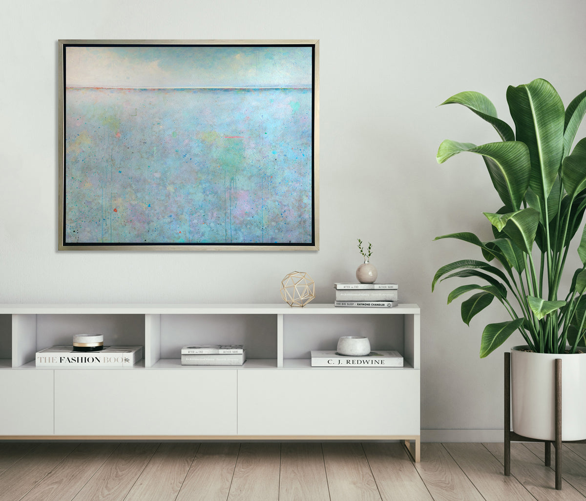 A blue abstract landscape print in a silver floater frame hangs on a white wall above a white console shelf with decorative accessories and a large potted floor plant.