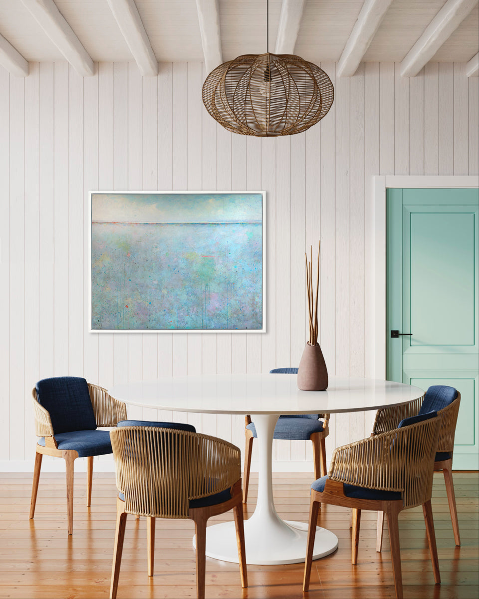A blue abstract landscape print in a white floater frame hangs on a white paneled wall next to a teal door and above a white pedestal table and five rattan chairs with navy cushions.