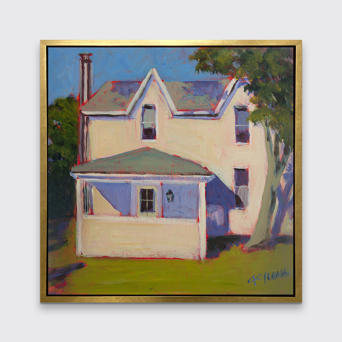 A print of a white house that sits next to a giant tree, green grass in front in a gold floater frame hangs on a white wall.