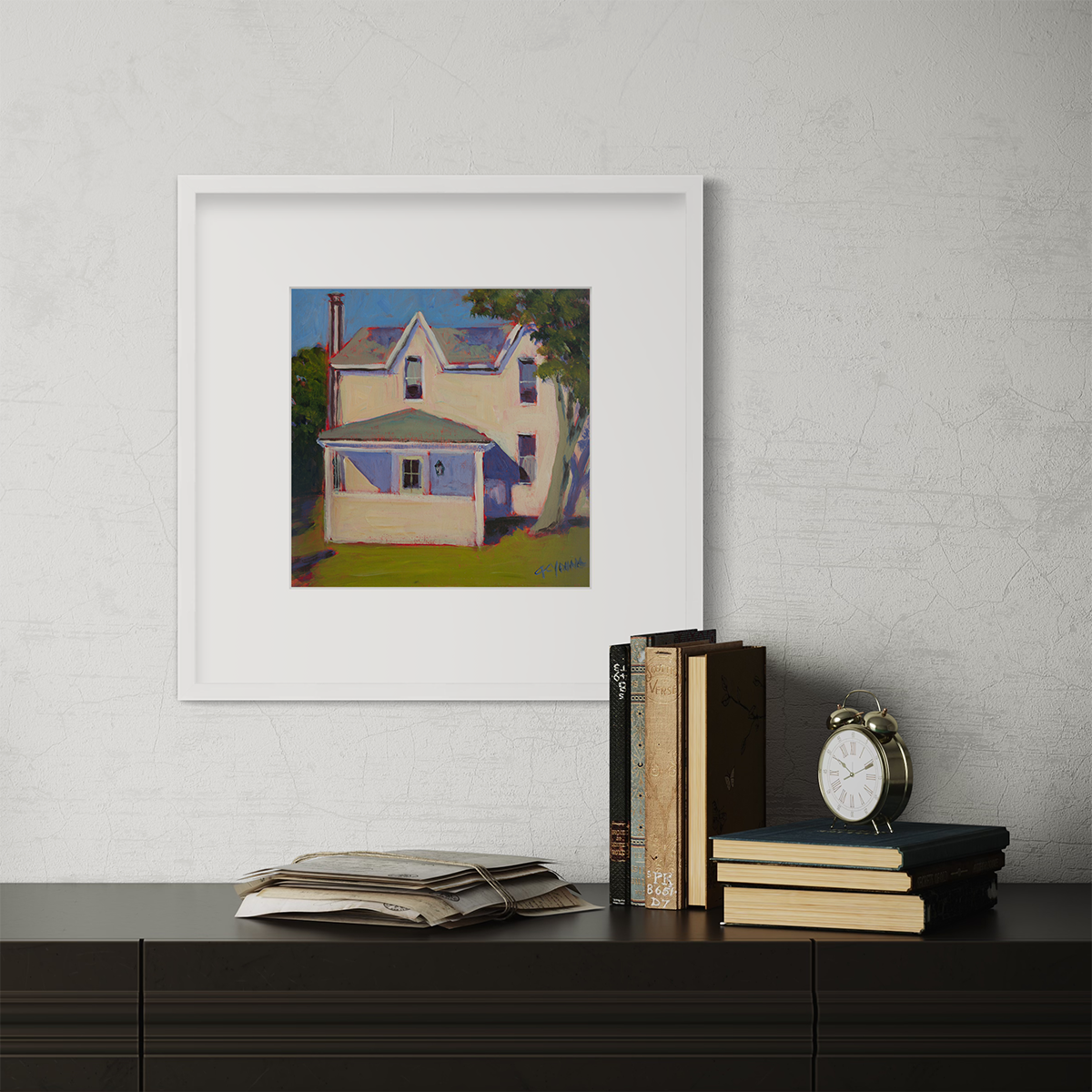 A print of a white house that sits next to a giant tree, green grass in front in a white frame with a mat hangs on a white wall above a dark surface with stacks of books and a small alarm clock.