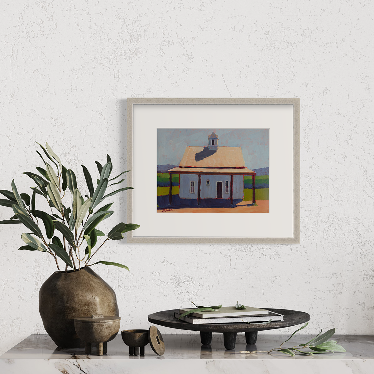 A contemporary house print in a silver frame with a mat hangs on a white wall above a white marble surface with a greenery filled vase and a small round platter.
