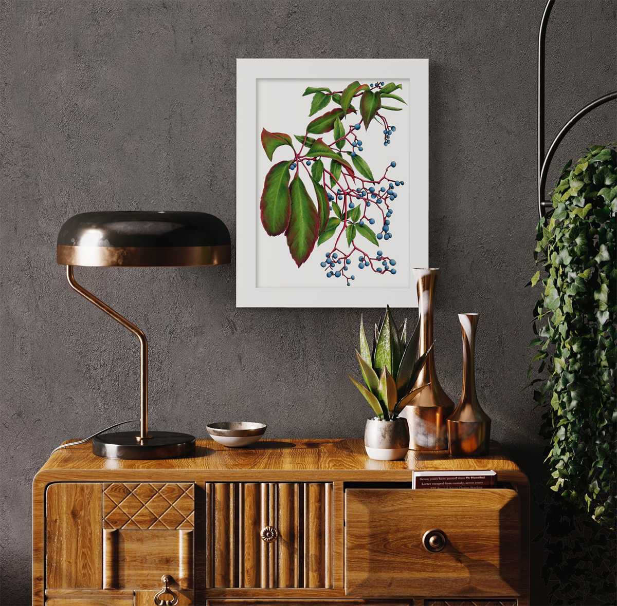 A green plant print with blue berries in a white frame hangs on a dark grey wall above a modern wood dresser with a lamp and decorative accessories.