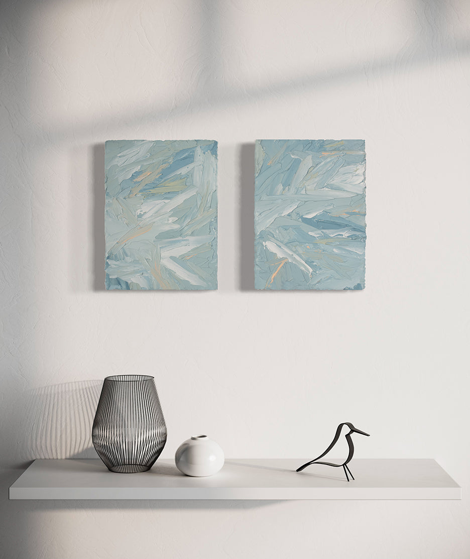 A pair of thickly painted paintings in teal, sea foam green, celadon, white and hints of orange and yellow by Teodora Guererra hang on a white wall over a white shelf with a metal bird sculpture, metal wire vase and white smaller ceramic vase sitting on it. The two painting are like wall sculptures.