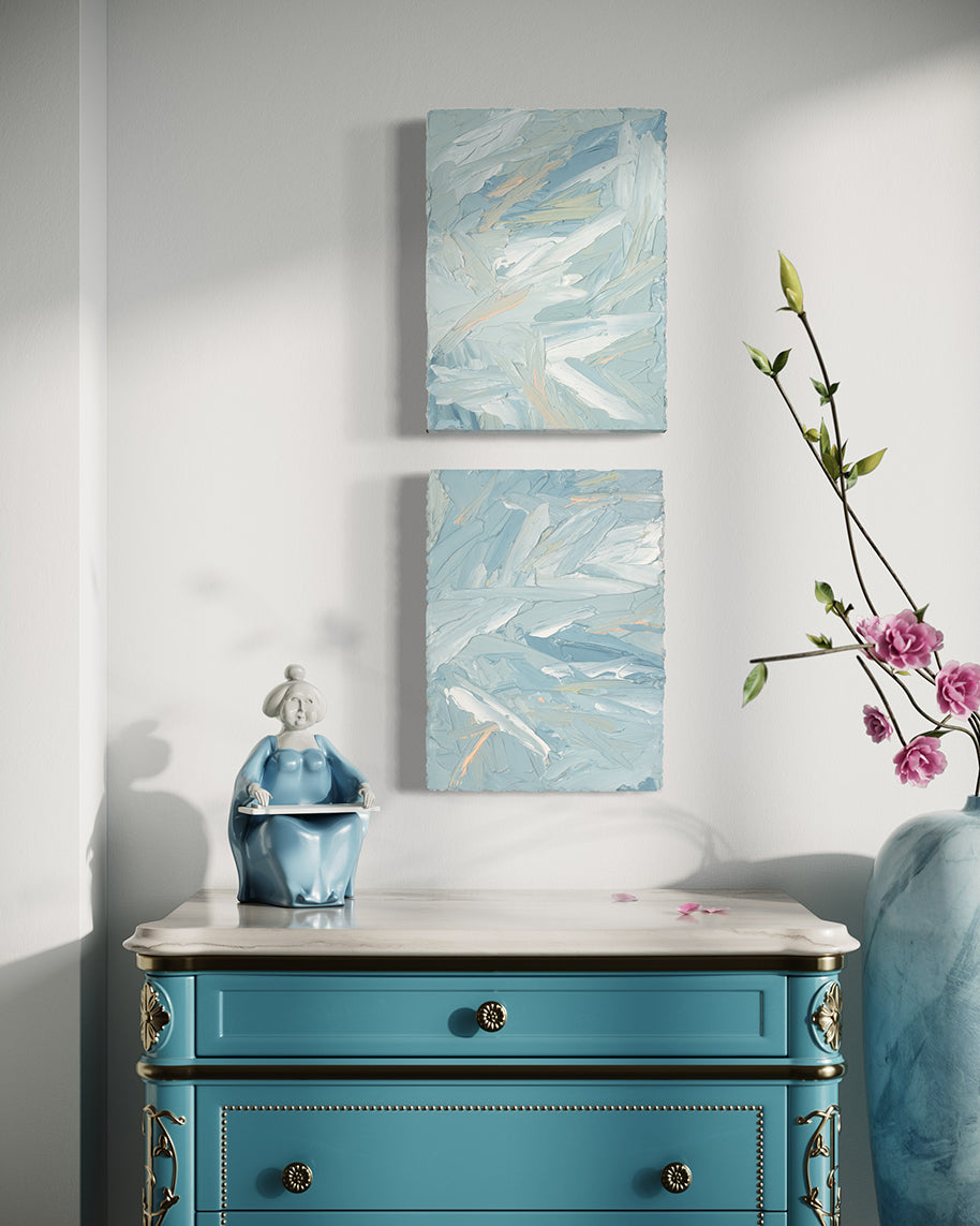 A pair of thickly painted paintings in teal, sea foam green, celadon, white and hints of orange and yellow by Teodora Guererra hang on a white wall over an ornate teal end table with white marble top. To the right is a marble vase with pink flowers and sitting on the table is a figurine of an asian woman in a teal dress. The two painting are like wall sculptures.