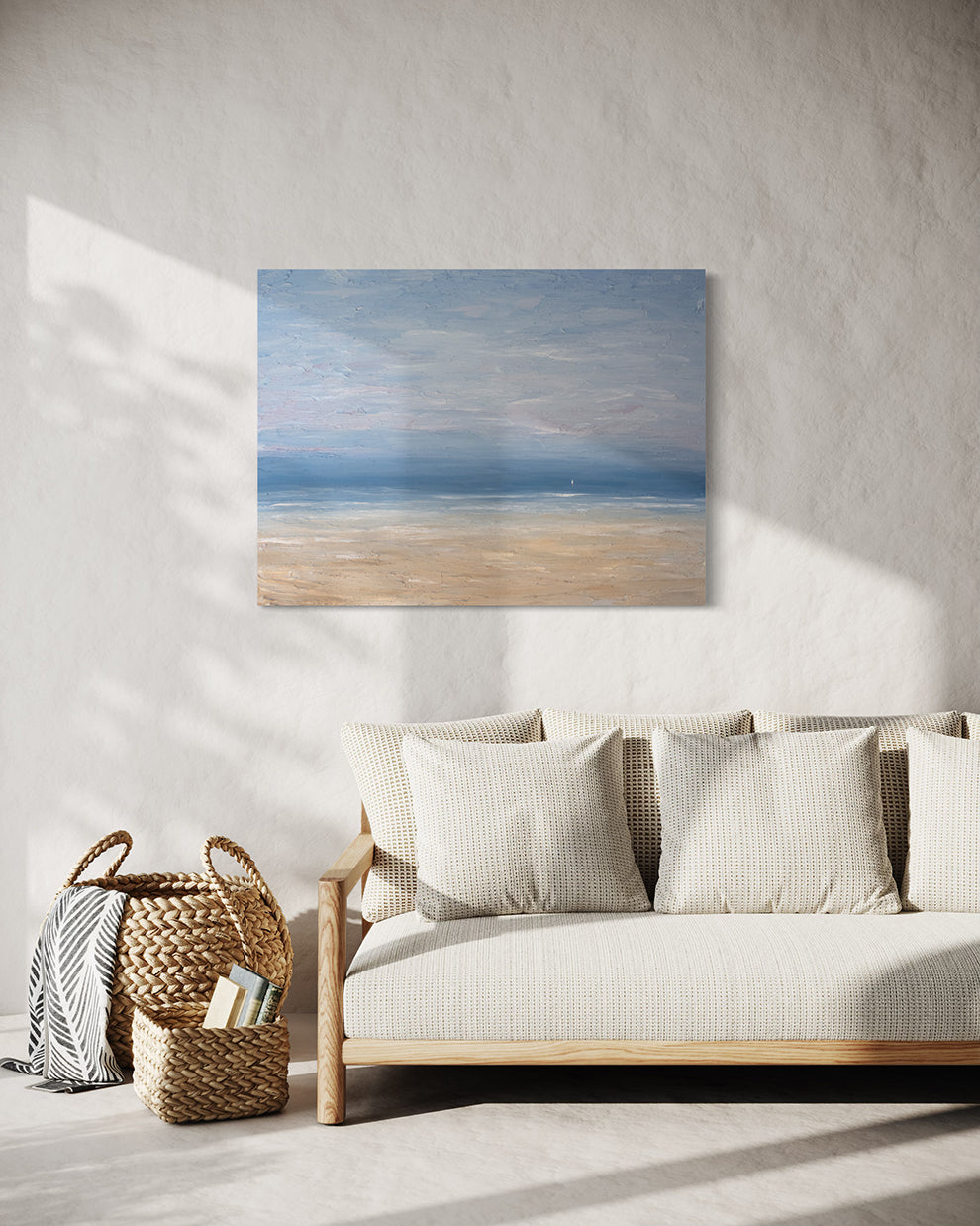 A very textured coastal painting on a white wall over a cream sofa with 2 baskets with books and a throw to the left.