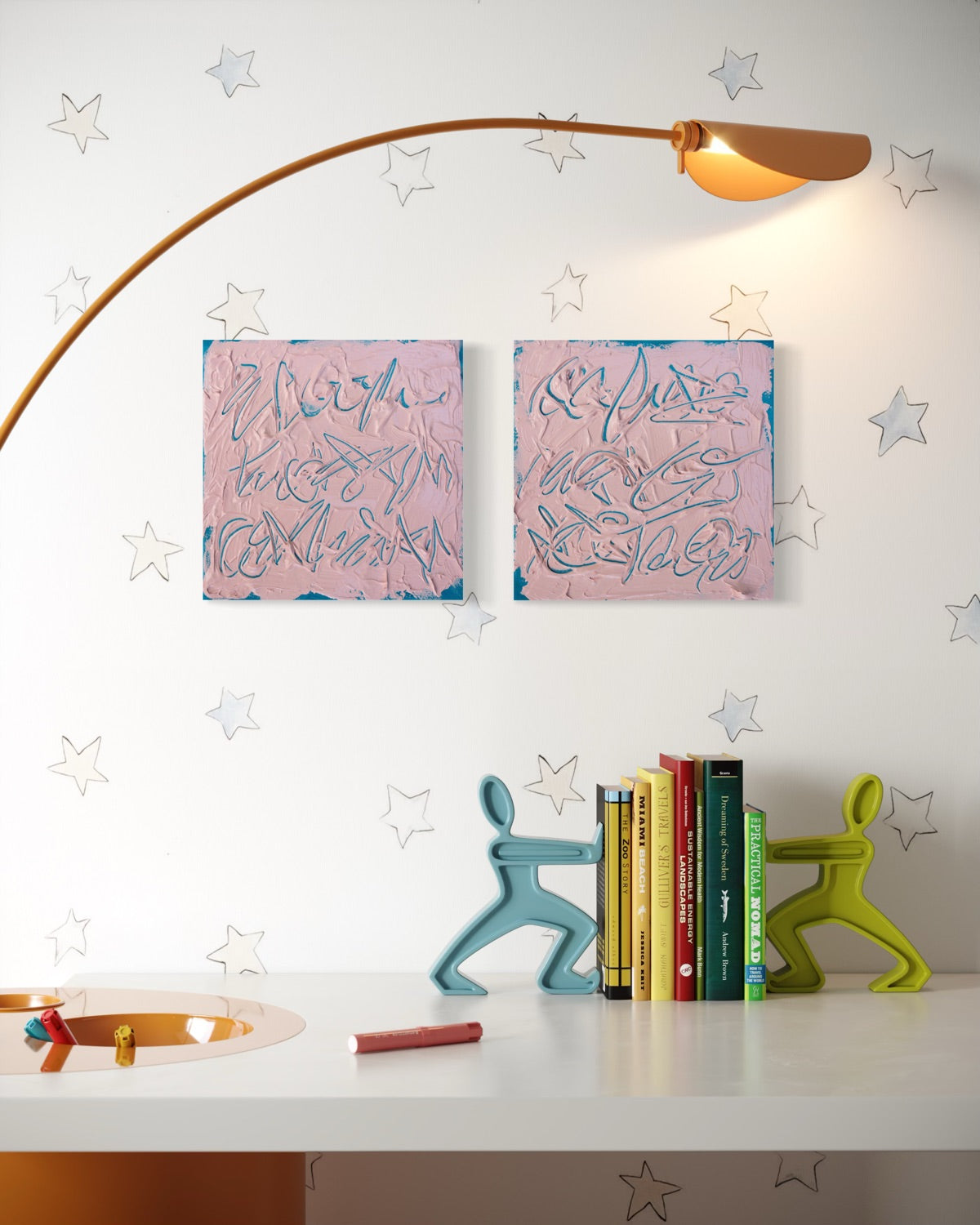 A styled interior scene with two pink abstract texted based paintings hanging on a white wall, a curved brass floor lamp, decorative star decals, a white table, books, and two colorful figurine bookends.