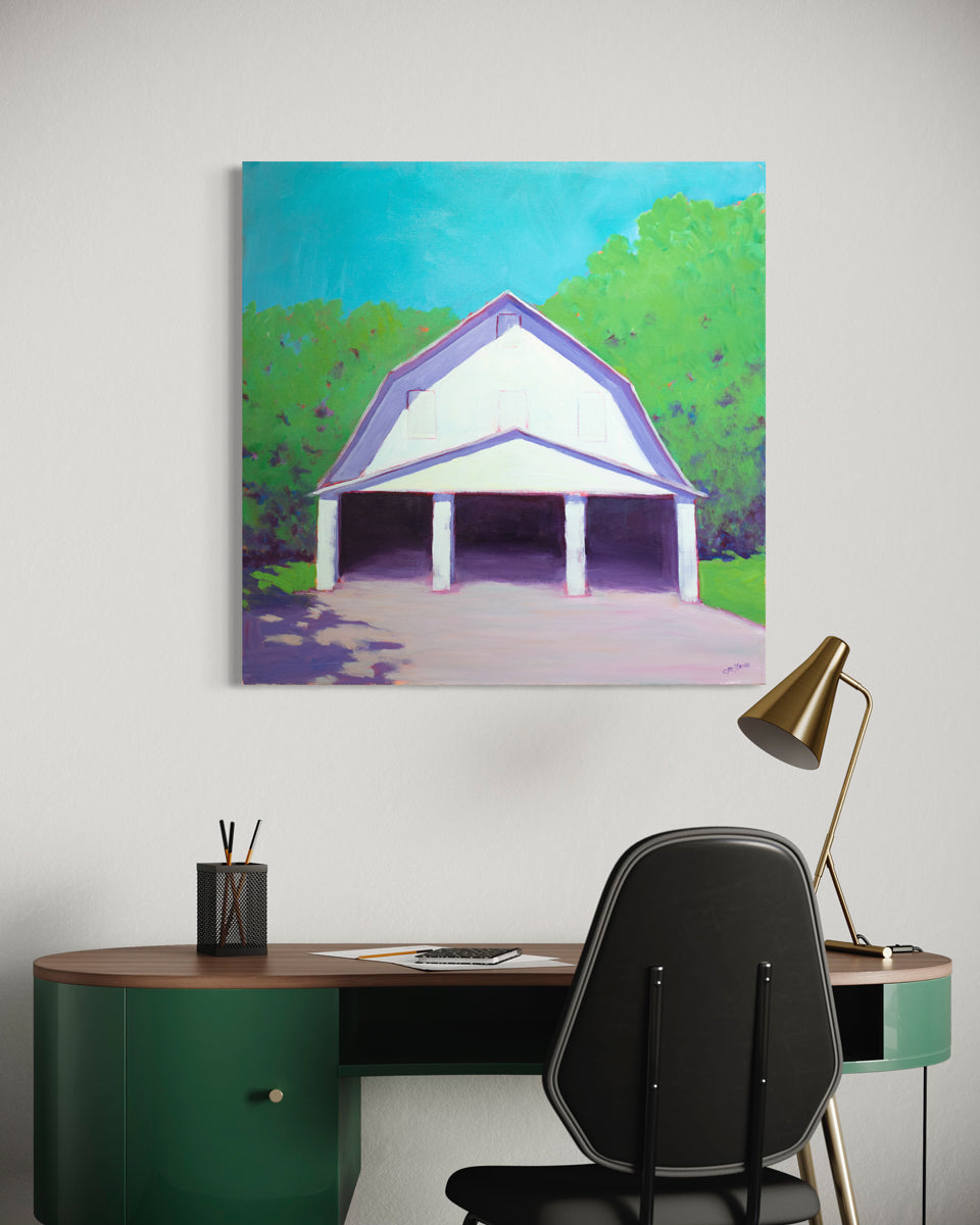 A landscape painting of a white barn with three open doors, lavender shadows, blue sky and surrounded by greenery by Carol Young hangs on a white wall above a green desk and a black chair.