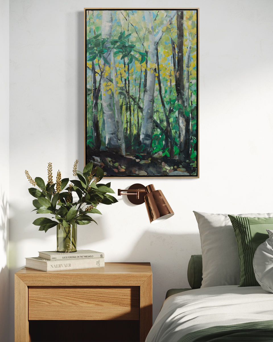 A green painting of a forest hangs on a white wall above a wood end table with books and plants in a vase, a floating wall lamp and a bed with green and white sheets. 