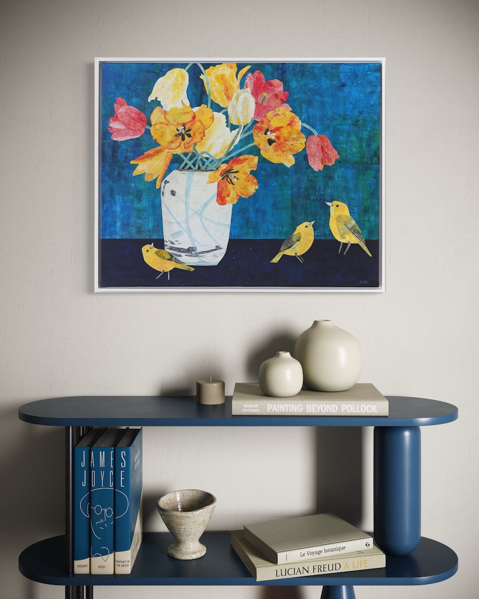 A framed floral collage with yellow, orange, and pink flowers on a dark blue background, displayed on a light gray wall above a blue shelf with books and ceramic vases.