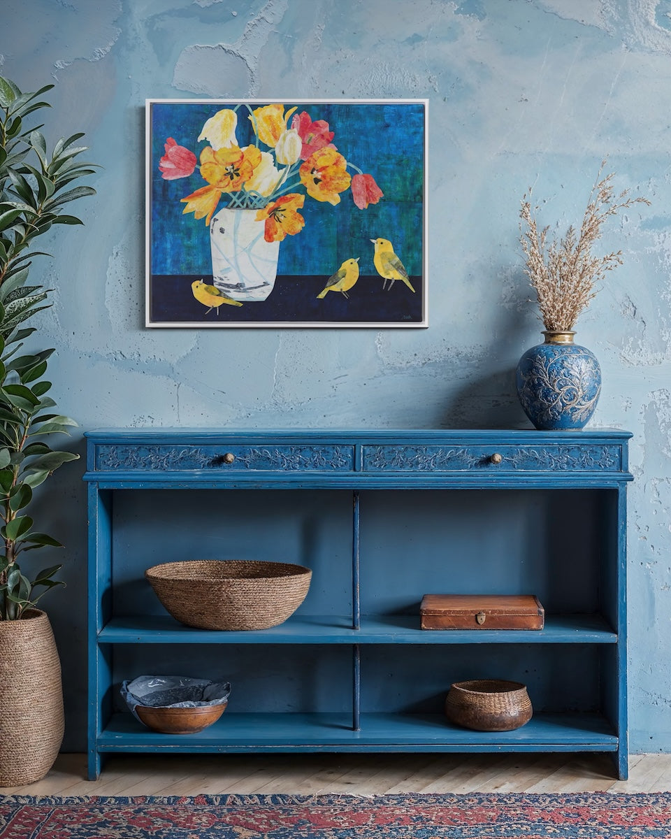 A framed collage depicting a vibrant bouquet of yellow, orange, and red flowers with small yellow birds on a dark blue background, displayed on a textured light blue wall above a blue wooden console table.