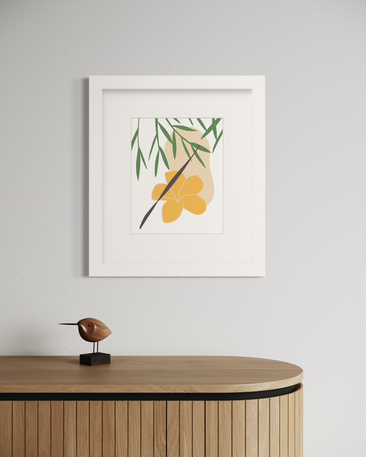 A green and yellow abstract botanical art print framed in a white frame hangs on a light wall above a wood surface with a small bird figurine.