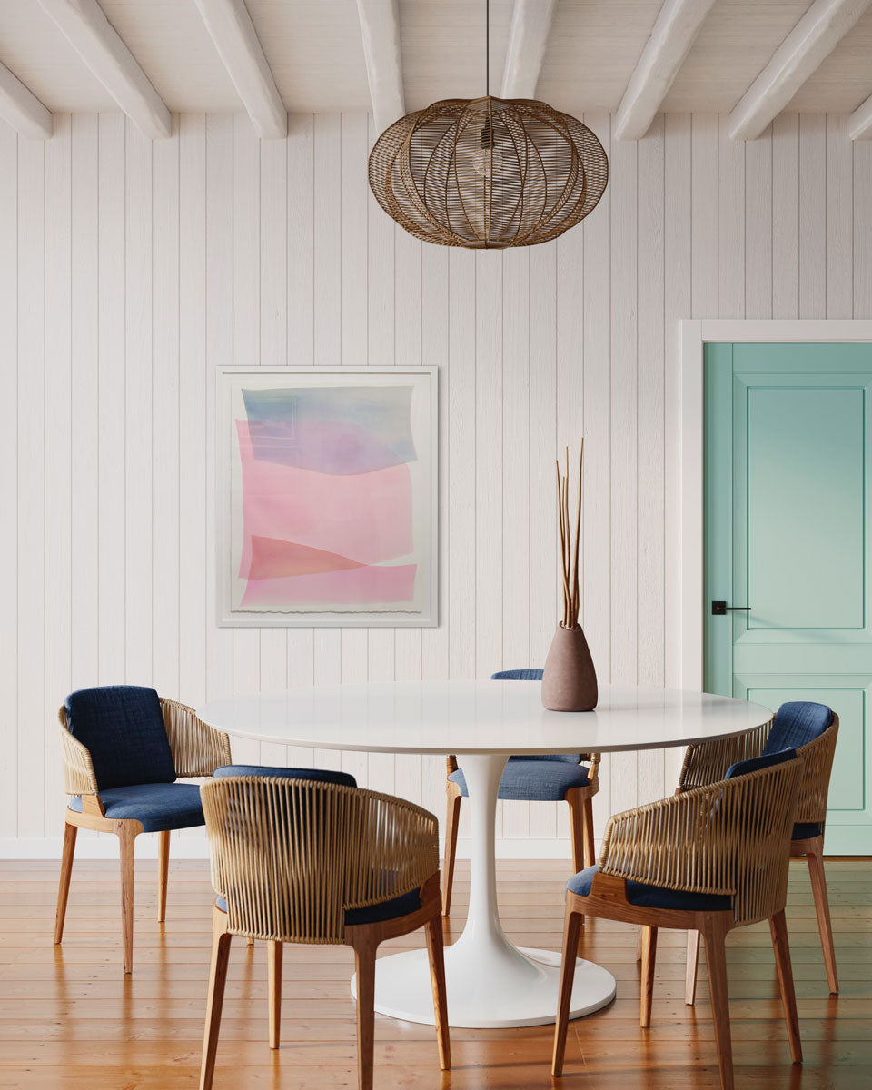 An abstract pink and blue watercolor painting framed in a white frame hangs on a white paneled wall above a midcentury white circular dining table and chairs with blue seat cushions. To the right of the painting is a light turquoise door. 